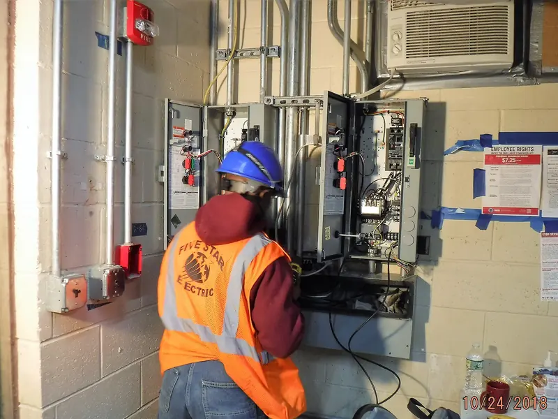 An electrician in a hard hat working at an open electrical panel