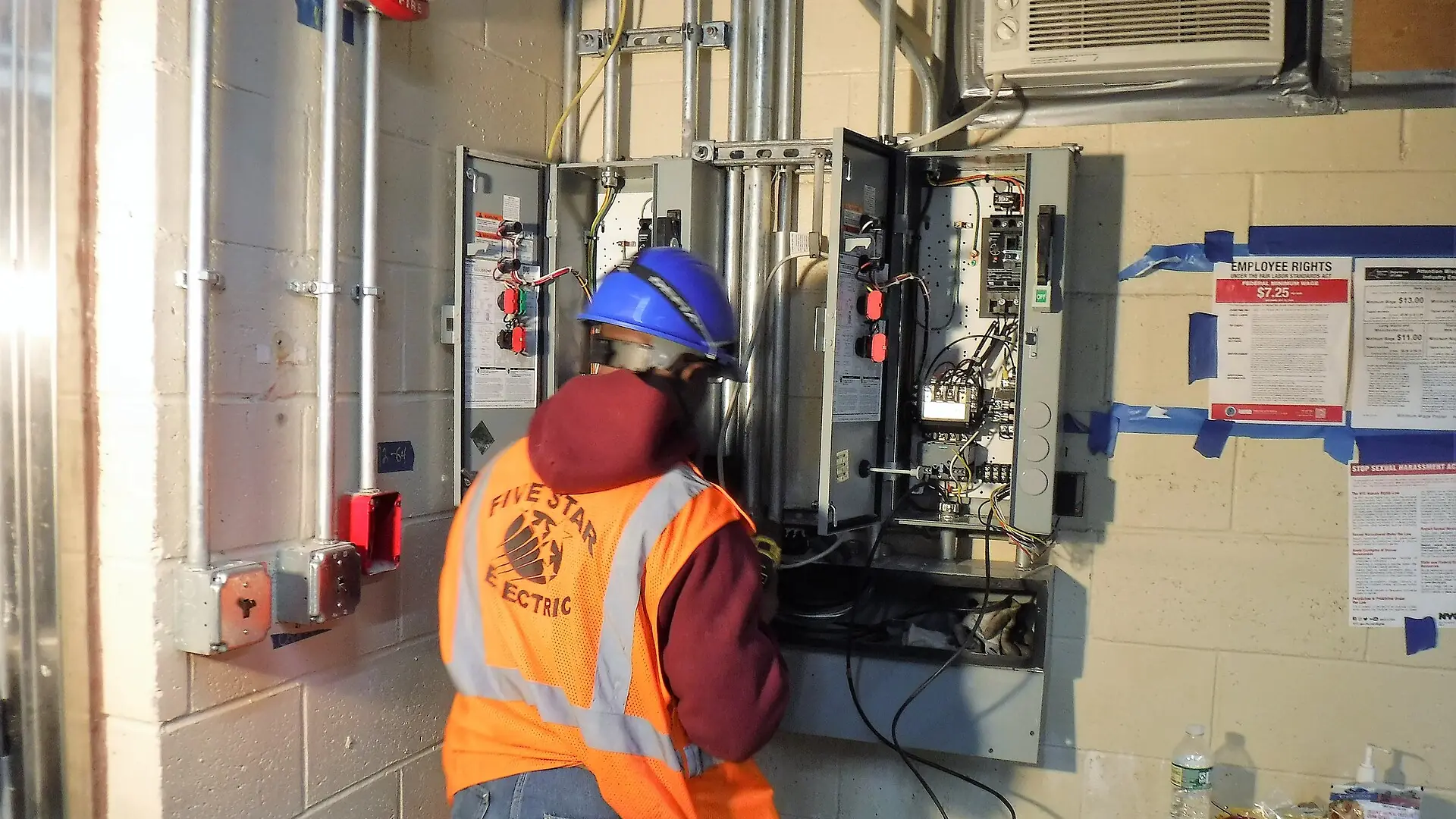 An electrician in a hard hat working at an open electrical panel