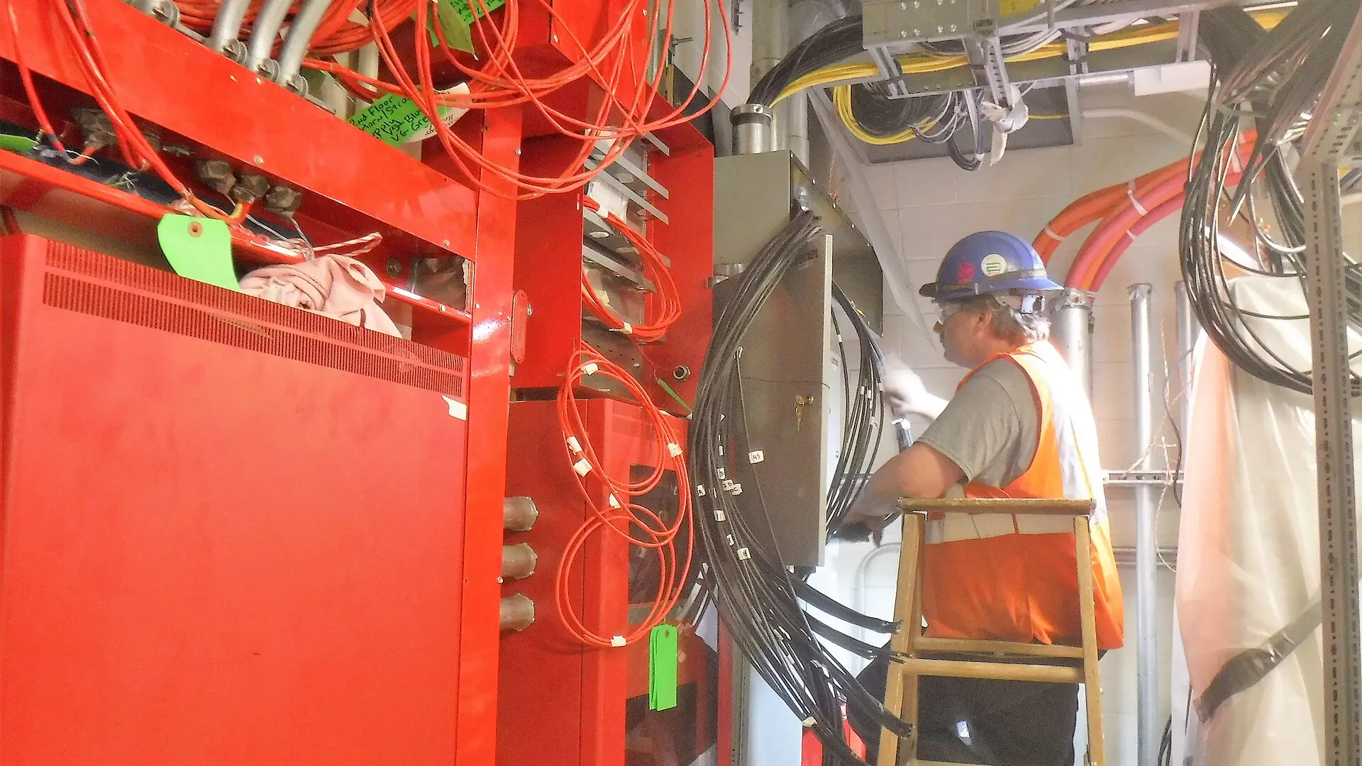 An electrician on a ladder pulling wire beside red fire-alarm equipment